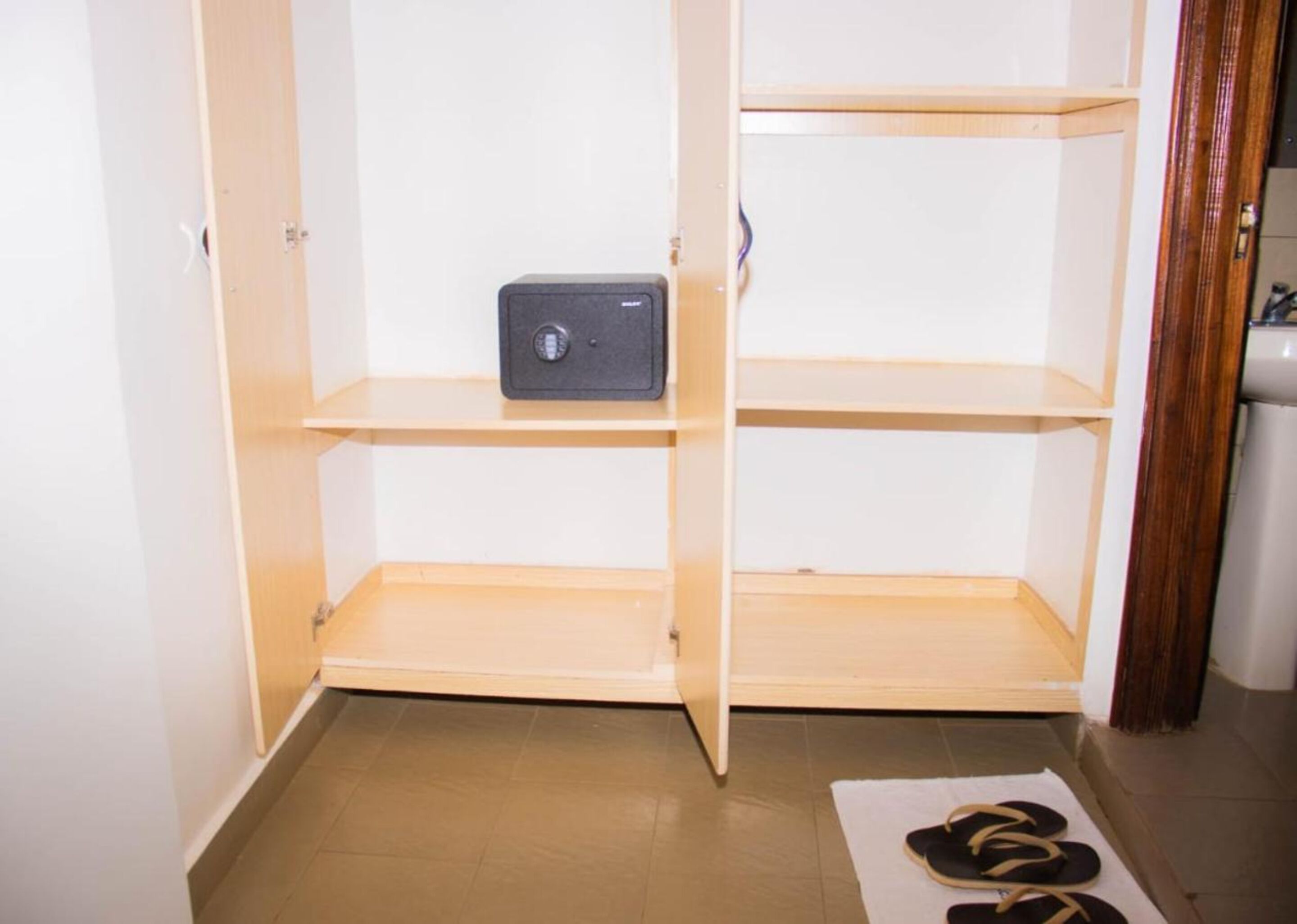A room with a wooden cabinet containing a safe, a pair of slippers on a mat, and a door leading to another room.