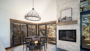 Un espace repas comprenant une grande table et des chaises, un lustre, une cheminée en pierre et de grandes fenêtres donnant sur une forêt.