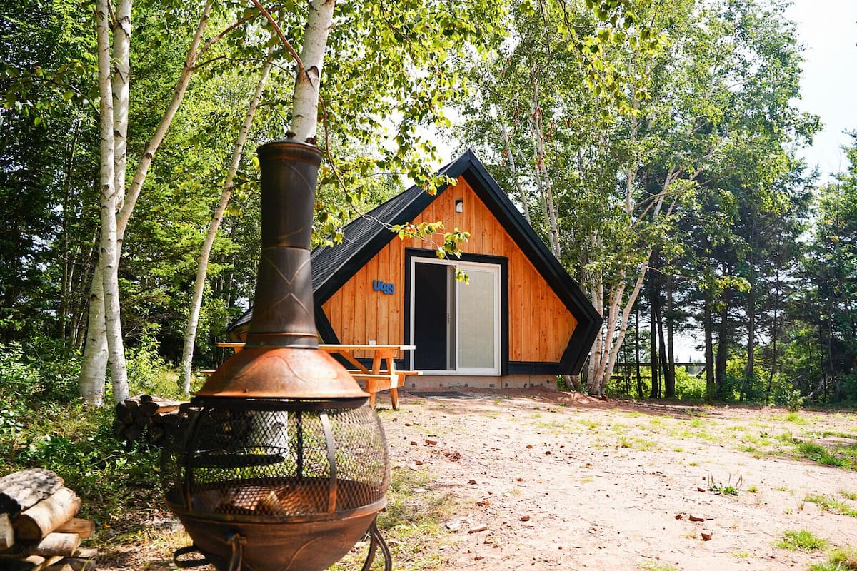 Uma cabana com uma chaminé grande, uma porta de madeira e uma mesa de piquenique em uma área arborizada.