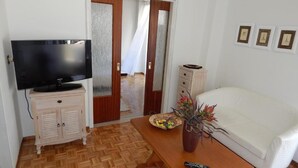 A room with a television on a cabinet, a white sofa, a wooden coffee table with a vase of flowers, and a door leading to another room. - Baroque 10-person Apartment in Makrygialos (Pydna-Kolindros)