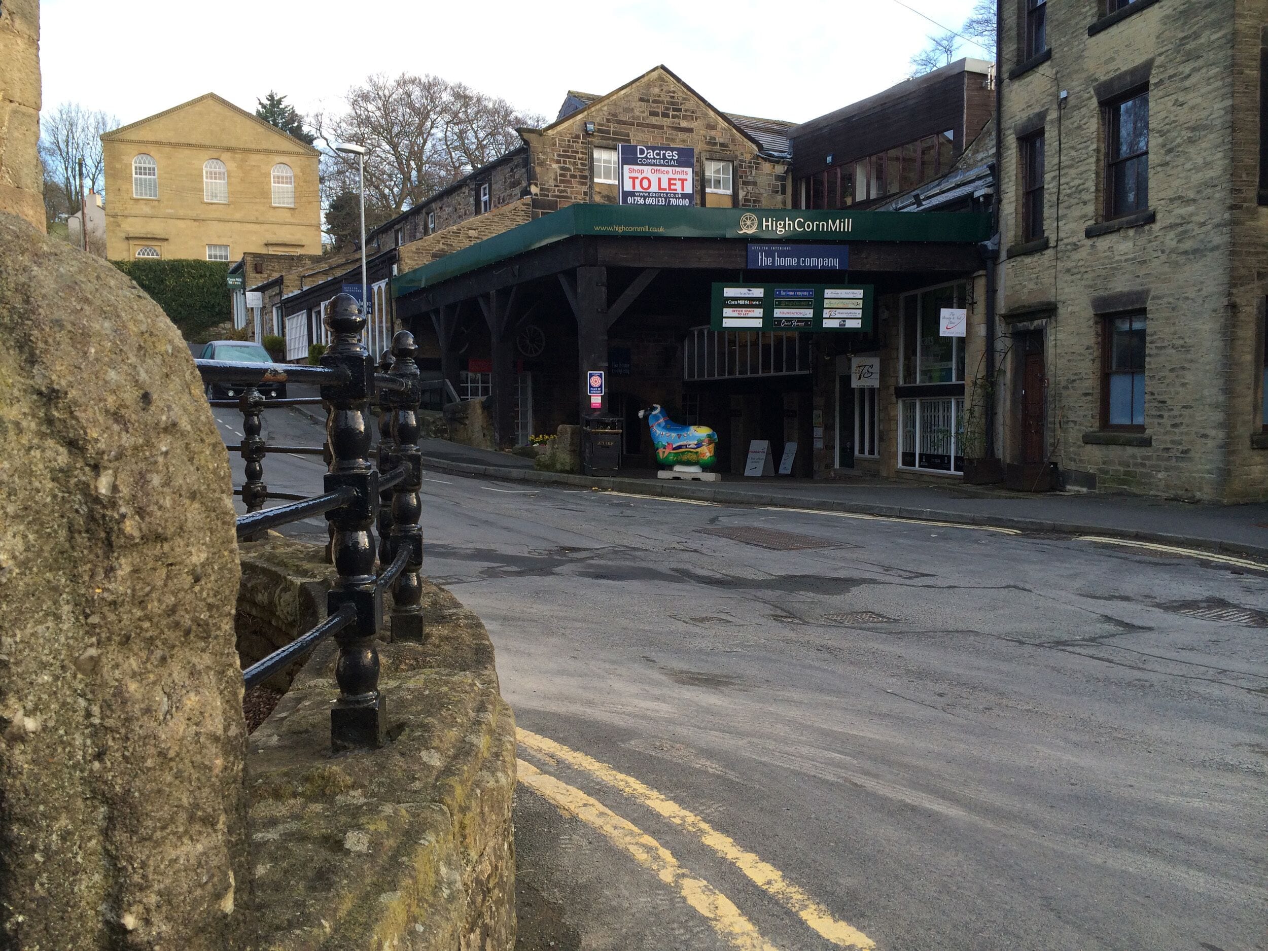 Calle con edificios, incluyendo un cartel que dice 'HighCornMill' y un anuncio de 'To Let'.