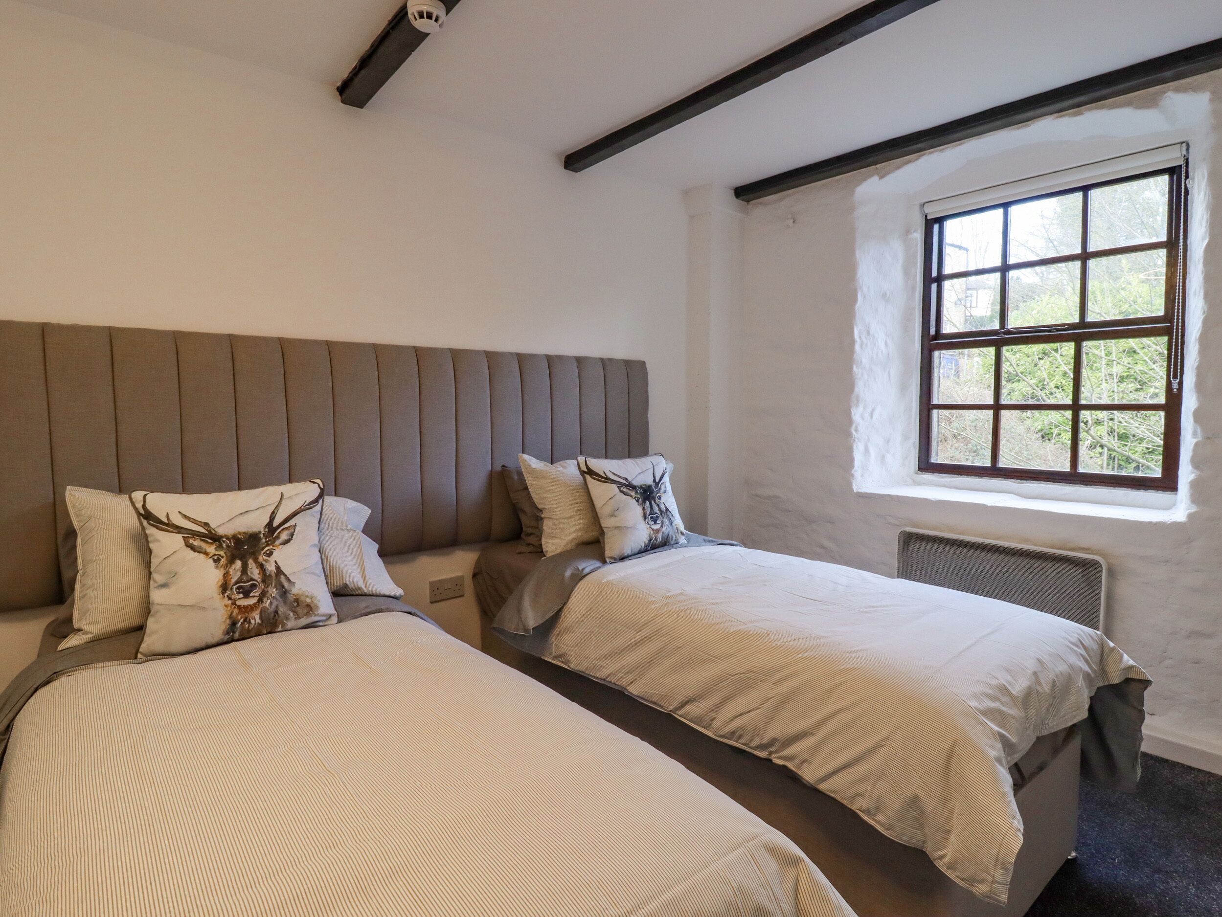 A room with two beds, a window with a view of greenery, and a ceiling vent.