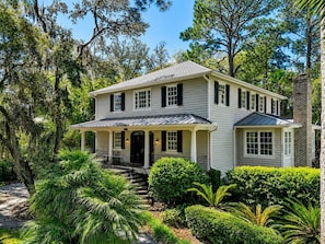 A two-story house with a porch, surrounded by lush greenery and trees. - 20 Canvasback: Beautiful Compound w/ Pool, 4th row (3 min walk) to the Ocean (Hilton Head Island)