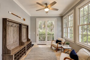 Quarto com um banco de madeira, ventilador no teto, porta com painéis de vidro e janela com vista para o verde.