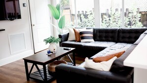 A modern living room with a black leather sofa, a wooden coffee table, and a large window with a view of greenery. - Cozy - full kitchen & 10ft island - 1.2G Wifi (Fort Lauderdale)