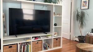 Ein Wohnzimmer mit einem großen Flachbildfernseher, weißen Regaleinheiten, Korbwannen und verschiedenen Dekorationsobjekten.