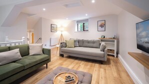 A spacious living room with a green sofa, a grey sofa, and a wooden coffee table. There is a floor lamp, a window with a view of trees, and a television on a wooden shelf. - Spacious Detached property with Waterfall View (Ambleside)