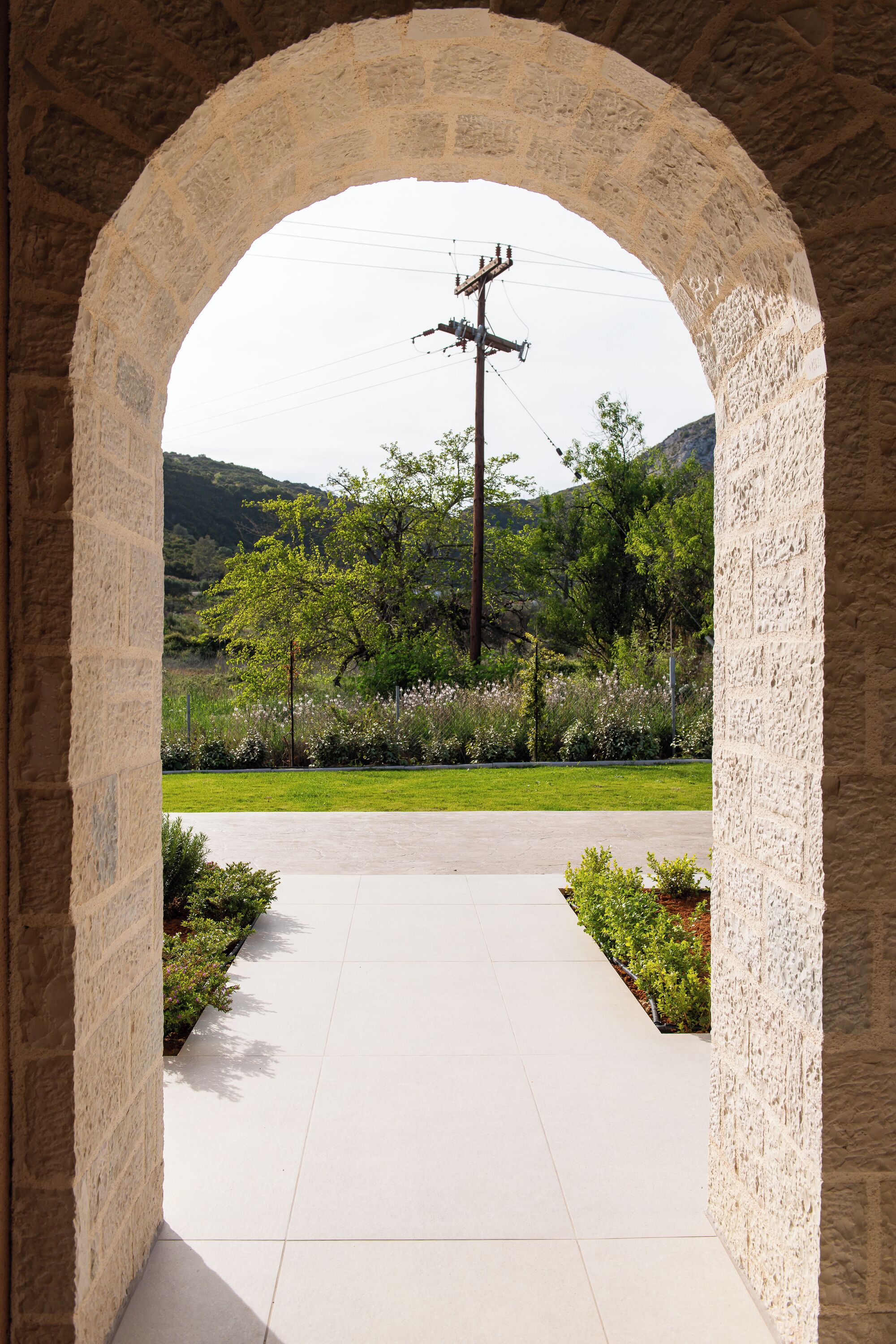 An arched stone doorway leading to a paved pathway with a view of a green landscape and a utility pole.