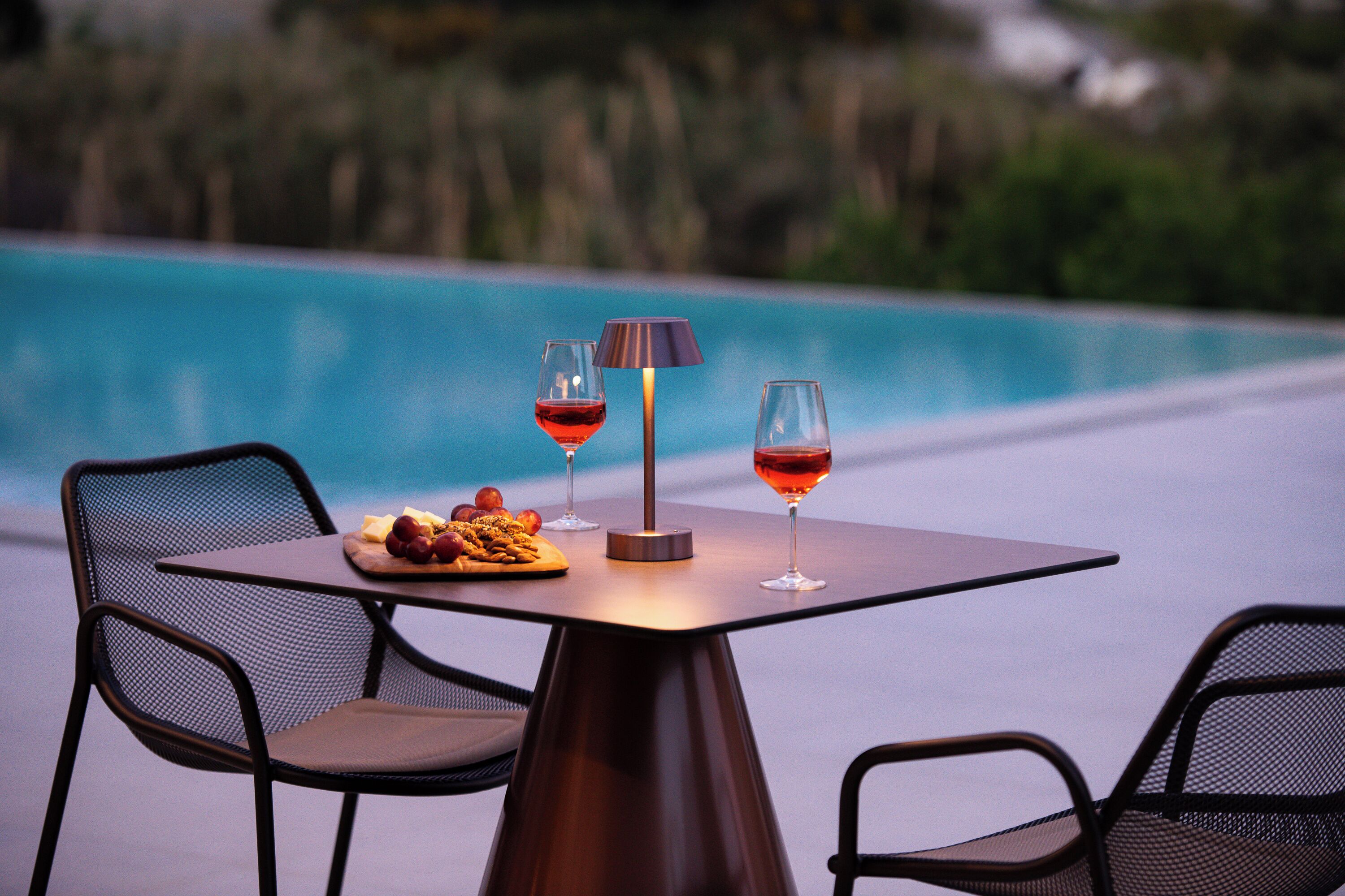 Mesa com duas cadeiras, abajur e dois copos de vinho tinto, próxima a uma piscina.