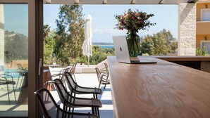 Un espacio exterior moderno para comer con una computadora portátil sobre una mesa de madera, rodeado de sillas y vistas a una piscina y follaje.