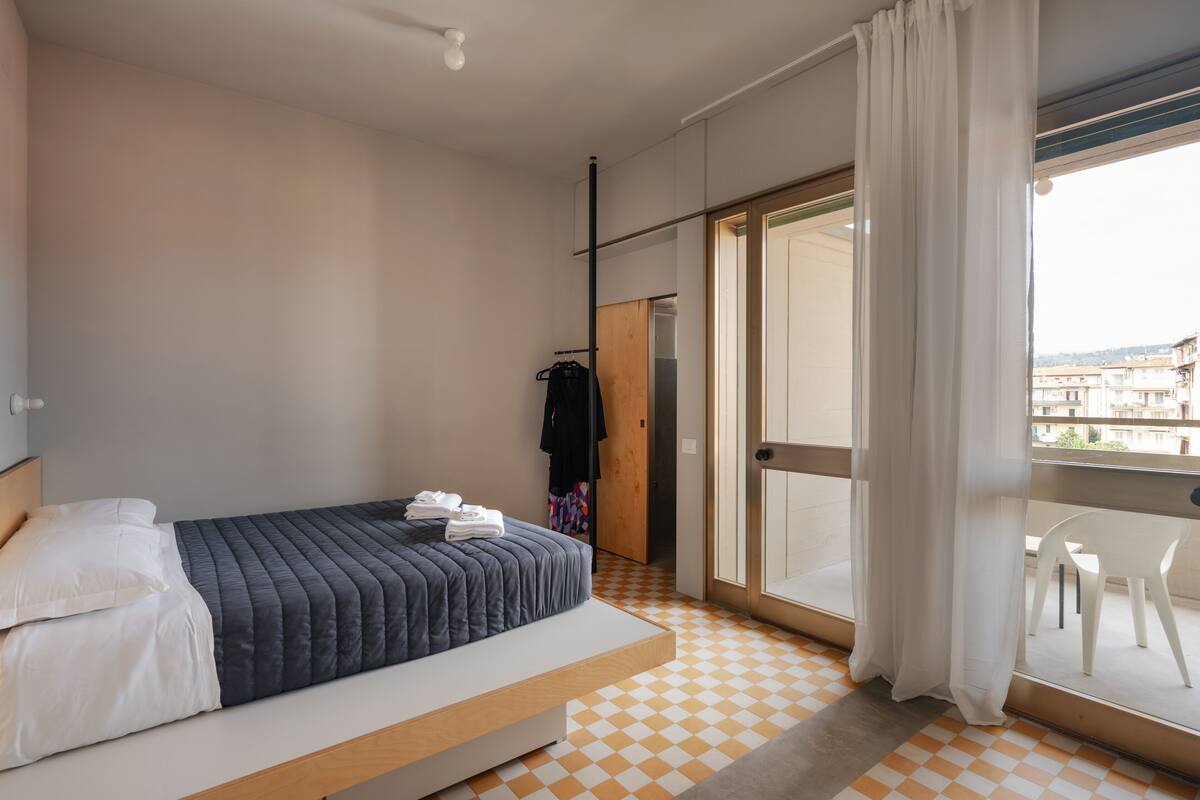 A bedroom with a bed, a wardrobe, a balcony with a chair, and a view of buildings.