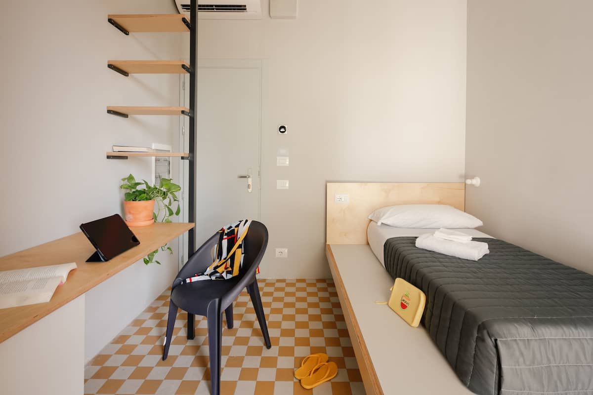 A compact hotel room with a bed, a desk with a chair, a potted plant, and a checkered floor.