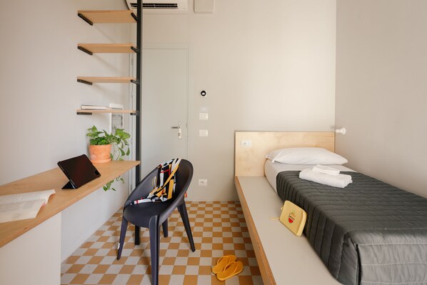 Une chambre d’hôtel de taille modeste, comprenant un lit, un bureau avec une chaise, une plante en pot et un sol à carreaux.