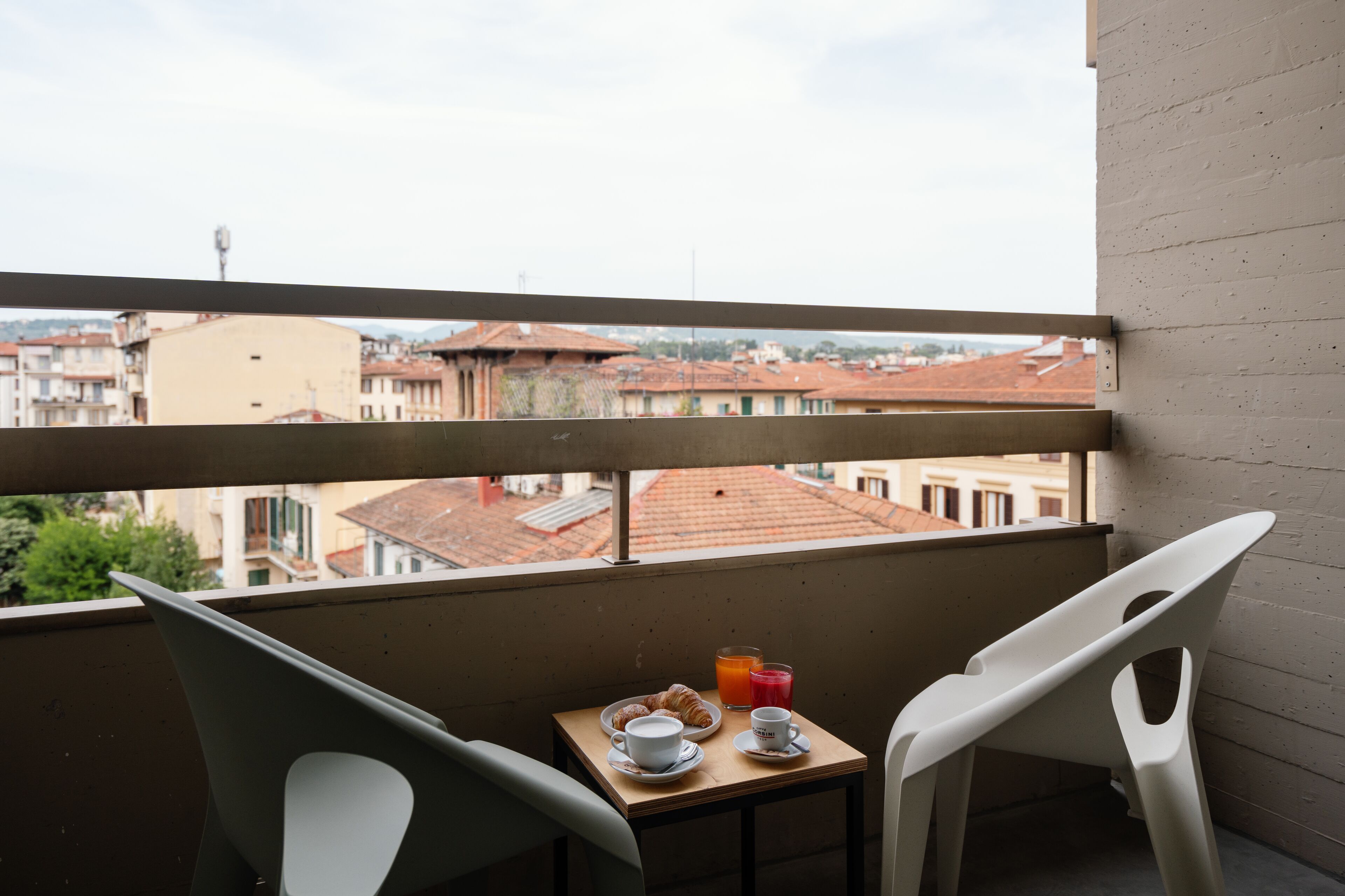 A balcony with a table set for breakfast, including croissants and juice, overlooking a cityscape with red-roofed buildings.