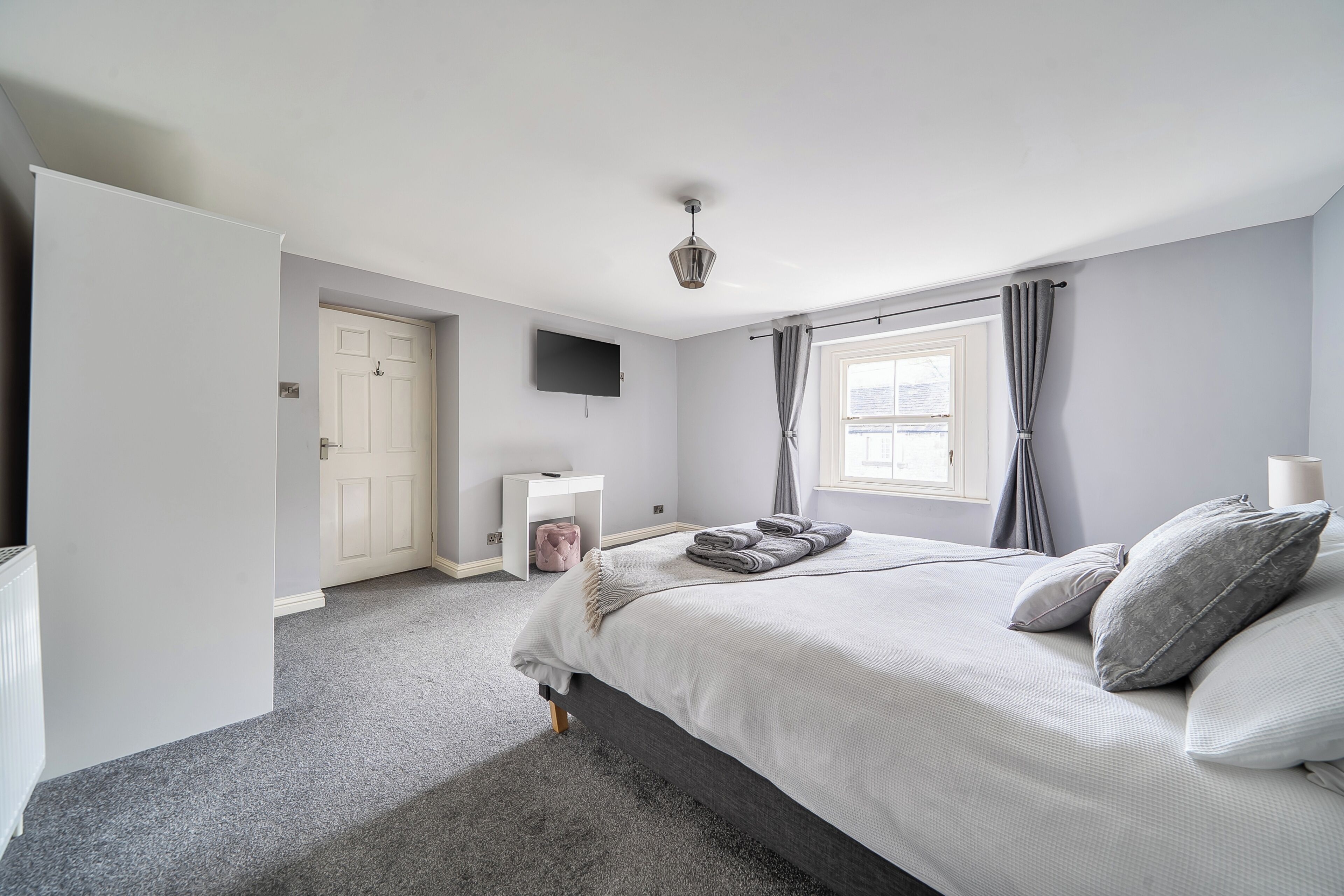 A bedroom with a bed, a bedside table, a TV, a window with curtains, and a door.