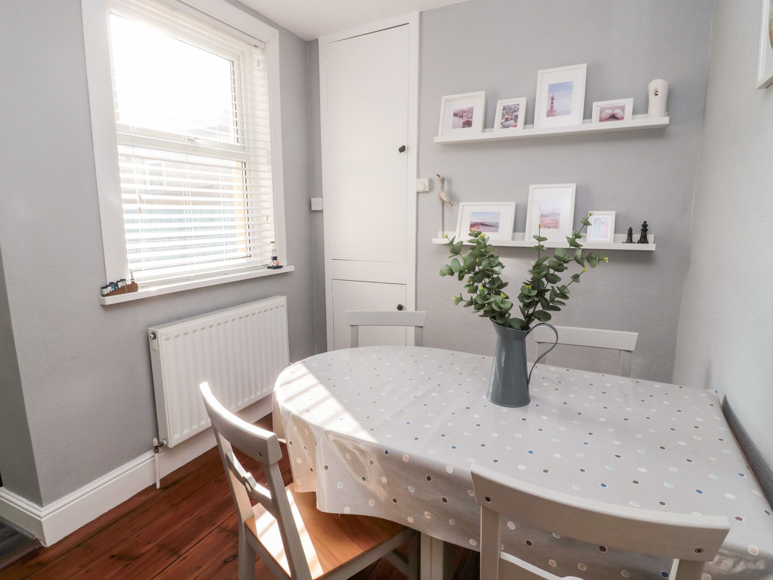A dining area with a round table, chairs, a vase with green plants, and a window with blinds.