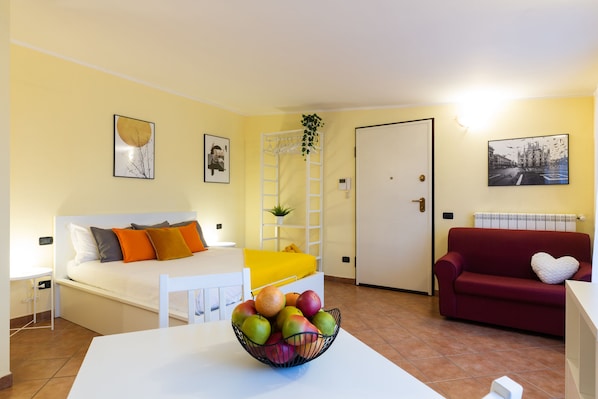 A room with a bed, a red sofa, a white table with a fruit bowl, and a white chair.