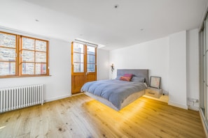 Quarto com uma cama grande, piso de madeira, radiador e janela com vista para um edifício de tijolos.