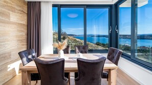A dining area with a table and chairs, large windows with a view of a coastal landscape. - Villa Astera -heated pool, 4 bedrooms, and sea views (Seget Vranjica)