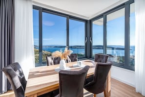 Un espace repas avec une table et des chaises en bois, de grandes fenêtres offrant une vue sur une étendue d’eau et sur la terre ferme au loin.