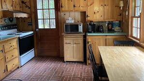 A wooden cabin kitchen with a stove, microwave, cabinets, and a window.