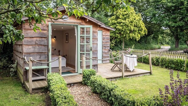 A rustic wooden cabin with a wooden deck, a bed, and a table set for dining. - Pebble Lodge, Toad Hall, Wangford (Beccles)