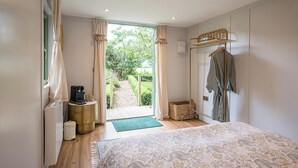 A bedroom with a bed, a bedside table with a lamp, a window with curtains, and a view of the garden. - Pebble Lodge, Toad Hall, Wangford (Beccles)