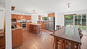 A spacious kitchen with wooden cabinets, a central island with stools, stainless steel appliances, and a large dining table.