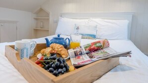 A neatly made breakfast tray on a bed with a white headboard, including croissants, grapes, strawberries, a croissant, a teapot, a glass of orange juice, and a newspaper.