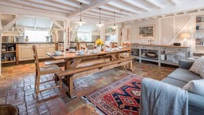 A spacious loft with exposed wooden beams, a dining area with a wooden table and benches, a kitchen with white cabinetry, and a cozy seating area with a patterned rug. - Ivy House Mells (Halesworth)