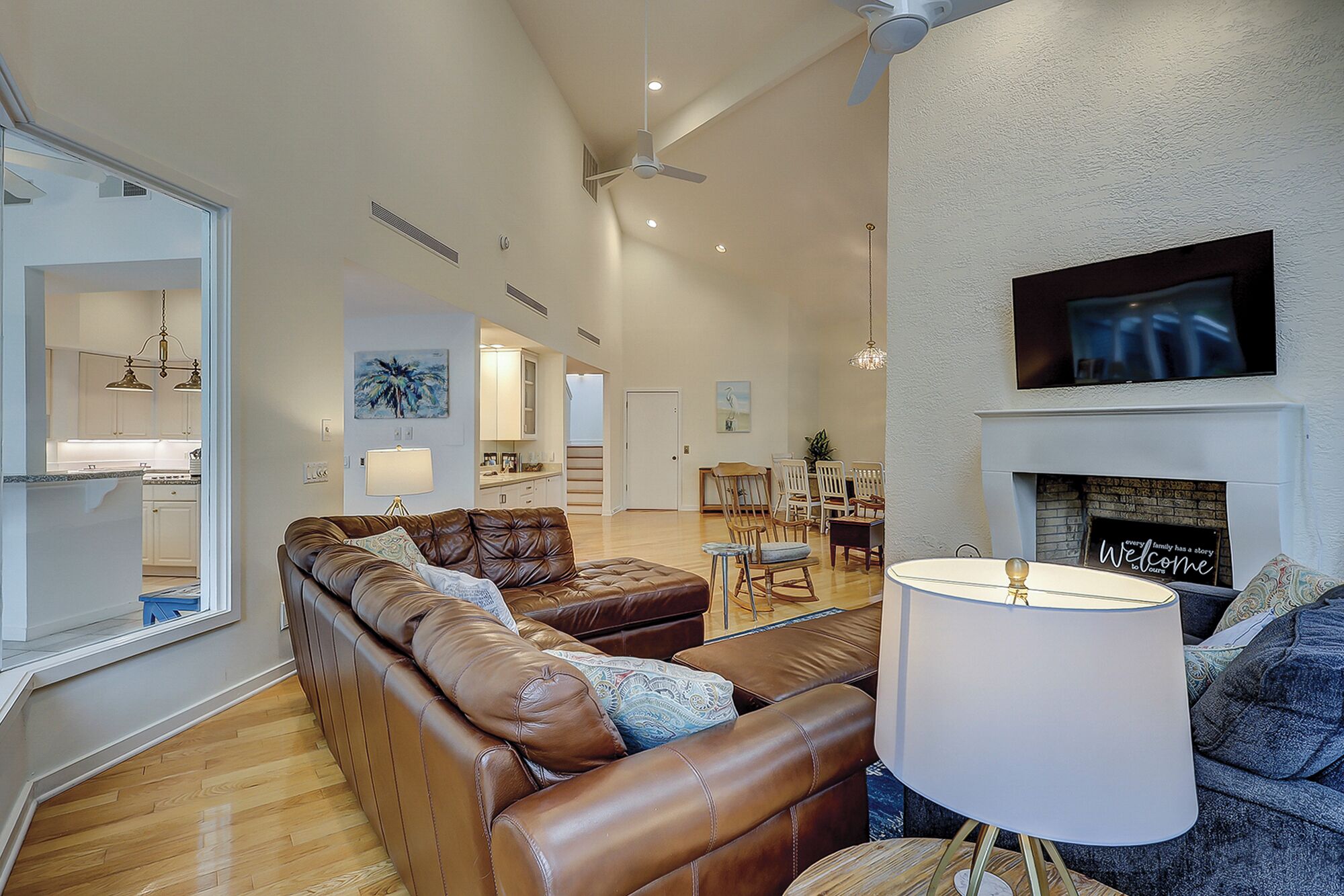 A spacious living room with a brown leather sofa, a dining area with wooden chairs, a fireplace, and a television mounted above it.