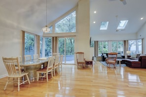 A spacious room with a wooden dining table, chairs, a sofa, and a rocking chair. Large windows provide natural light and views of greenery outside.