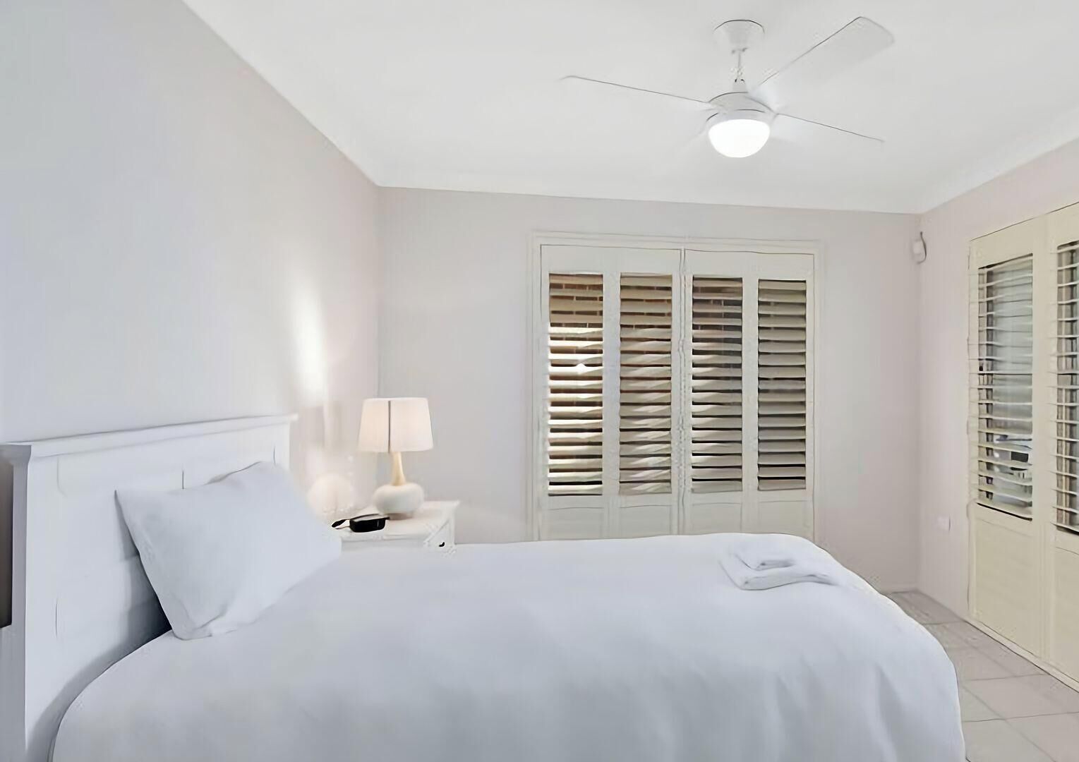 A bedroom with a bed, a nightstand, a lamp, a ceiling fan, and a window with shutters.