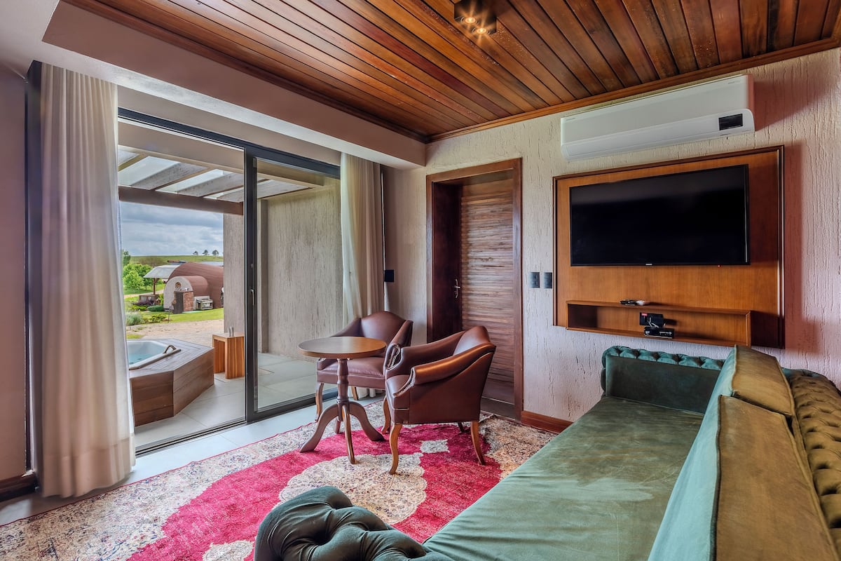 Una habitación moderna con un gran ventanal corredero que da a un área con jacuzzi exterior, un televisor de pantalla plana en la pared y una zona de estar acogedora con sofá y sillones.