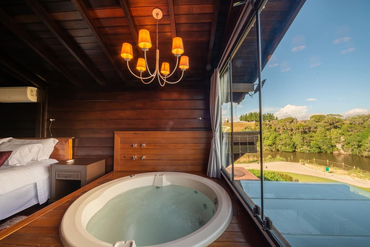 Una cabaña de madera con hidromasaje, una cama, una mesita de noche y vistas a un río.