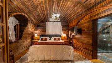 A cozy bedroom with wooden walls, a large bed with two pillows, a chandelier, and a view through a glass door.