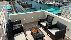 Una terrazza sul tetto con mobili in vimini, un tavolo con arance e bicchieri da vino, e una vista sul porto turistico sullo sfondo.
