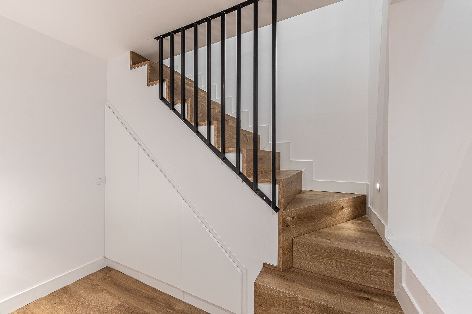 A staircase with wooden steps and black metal railings, leading to a landing with a white wall and a wooden floor.