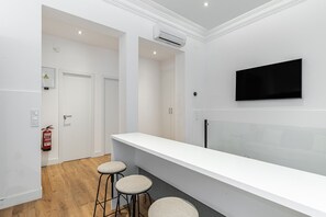 A modern hotel room with a white reception desk, two bar stools, a mounted TV, and a fire extinguisher.
