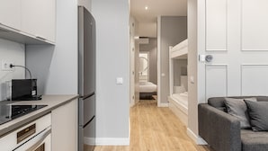 Une cuisine moderne équipée d’un réfrigérateur encastré, d’une machine à café et d’une horloge numérique. Un couloir mène à une salle de bain dotée d’un miroir et de toilettes. Un canapé gris est visible dans l’espace de vie.