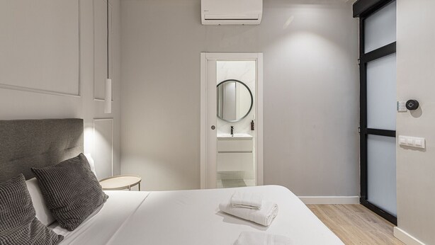 Une chambre moderne comprenant un lit, des oreillers, une table de chevet, un miroir, un système de climatisation intégré et une salle de bain avec un lavabo et un miroir.