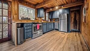 A cabin-style kitchen with wooden walls and a large window, featuring a stainless steel refrigerator, double ovens, and a sink.