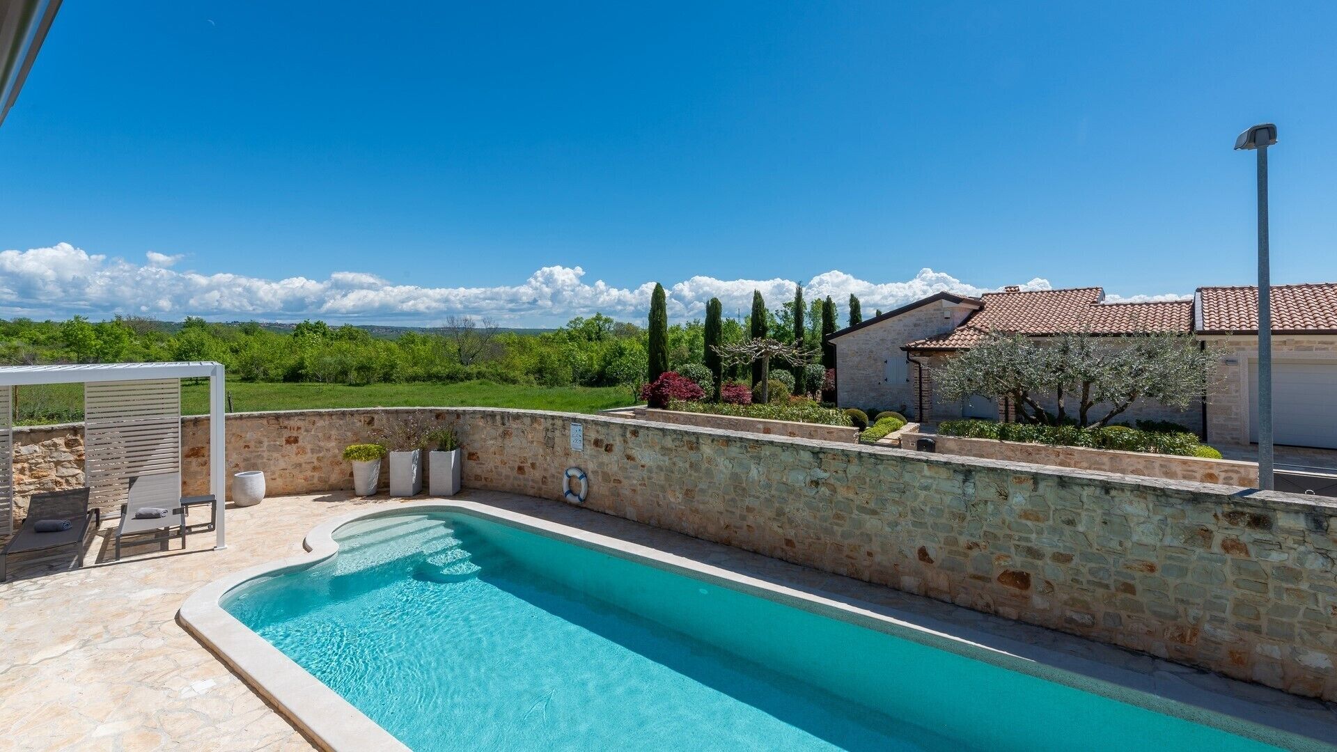 A swimming pool with a stone wall, a pergola, and a view of a house and landscape.