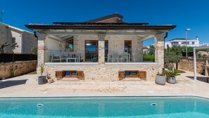 En moderne villa med pool, stenvæg og overdækket udendørs opholdsområde.