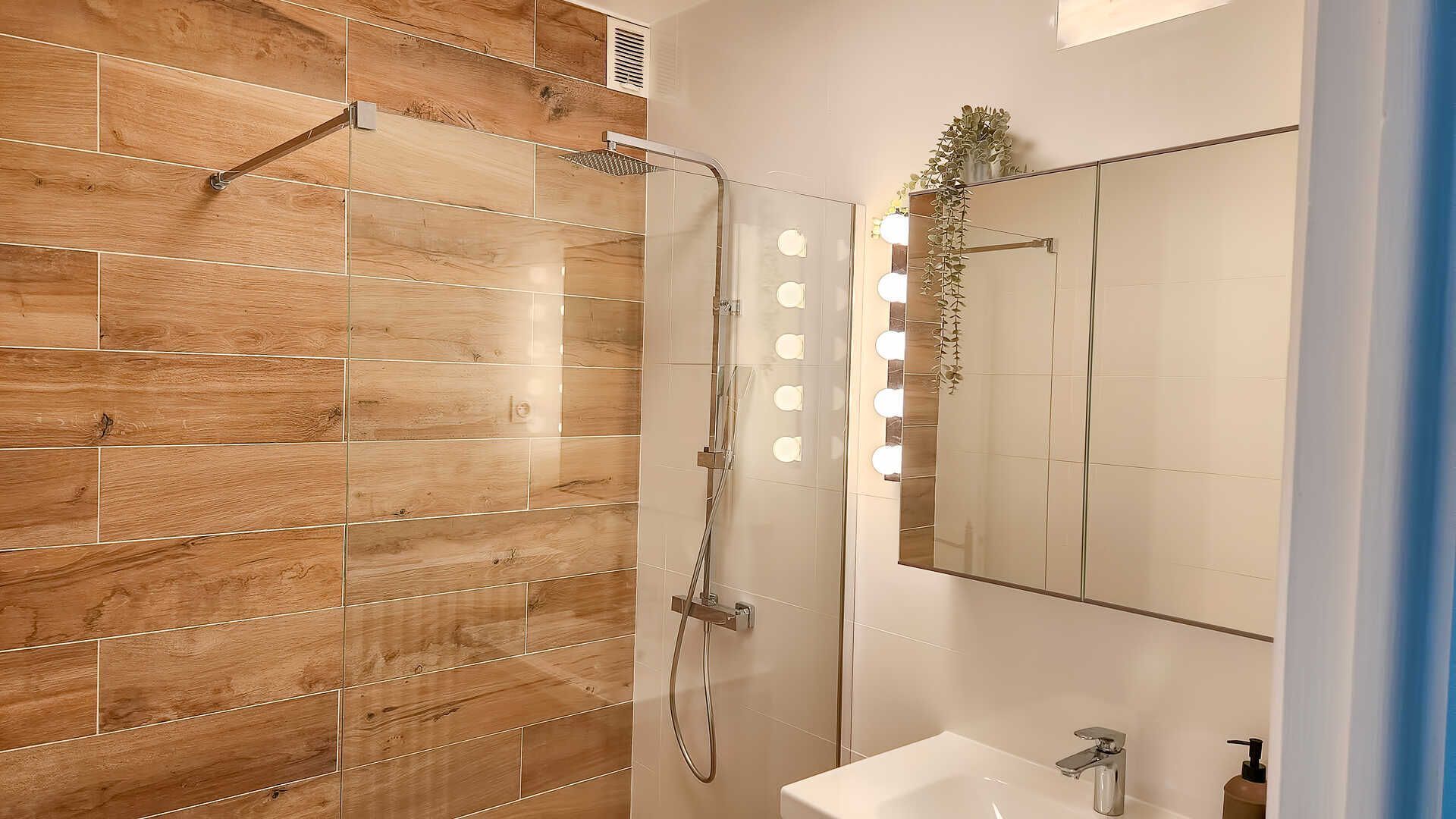 Un baño con pared de ducha revestida de madera, ducha con cerramiento de vidrio, espejo con iluminación y lavabo con dispensador de jabón.