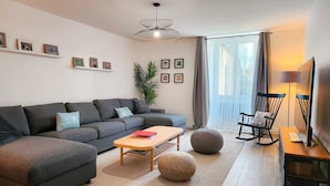 A modern living room with a grey sectional sofa, a wooden coffee table, a rocking chair, and a large window with curtains.