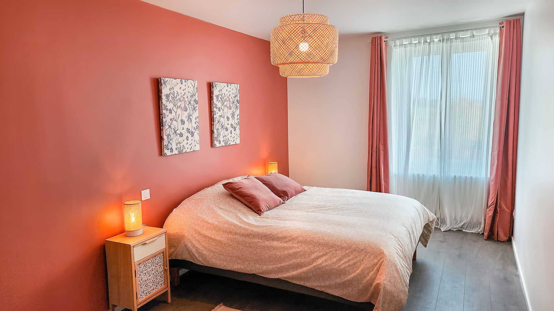 Una camera da letto con un letto, due opere d'arte appese al muro, un lampadario e un comodino con una lampada, oltre a una finestra con tende leggere.