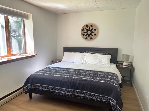 Un dormitorio con cama, ventana, un reloj de pared y mesita de noche con lámpara.