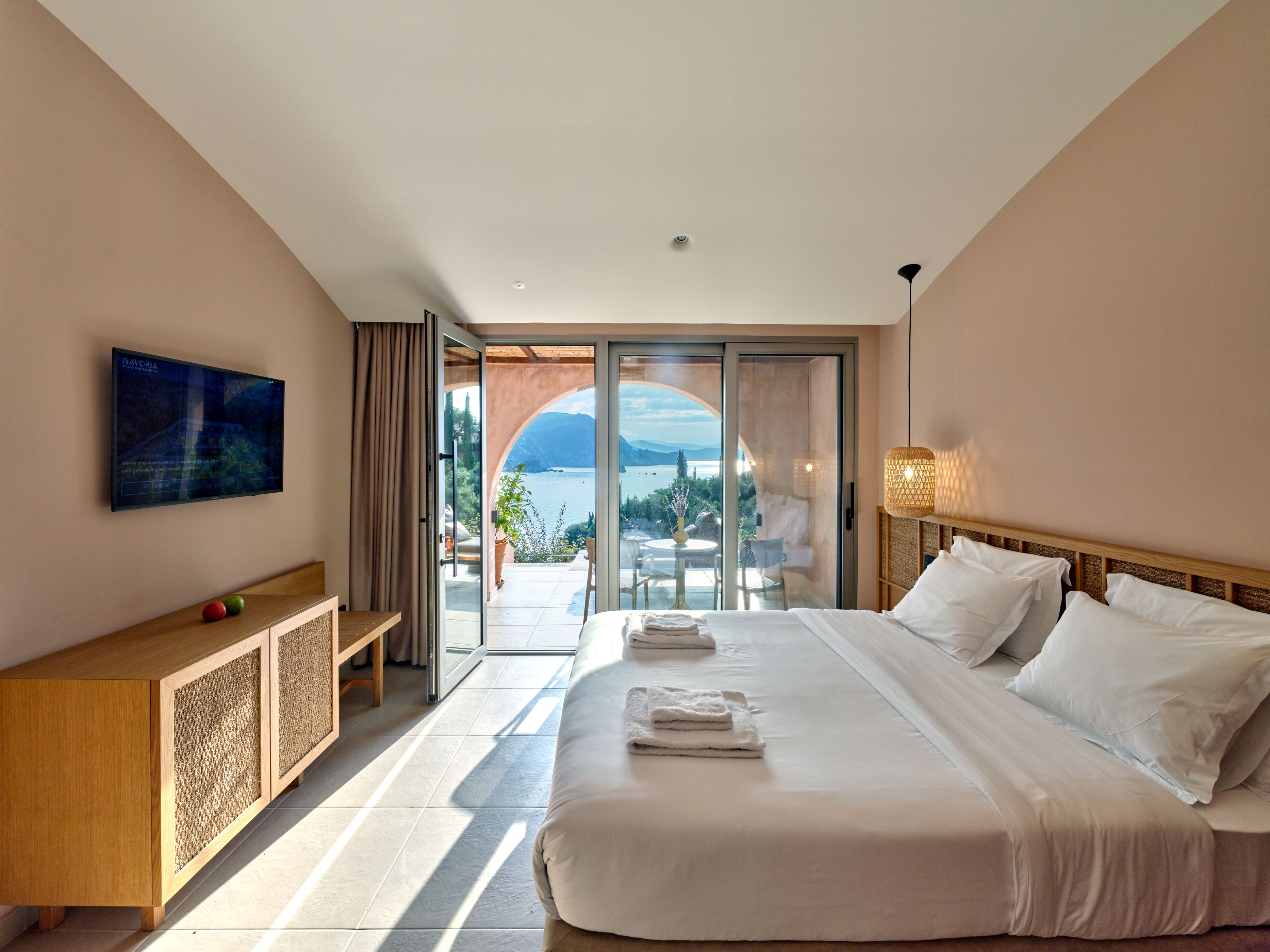 A hotel room with a large bed, a TV mounted on the wall, and a balcony with a view of the sea.