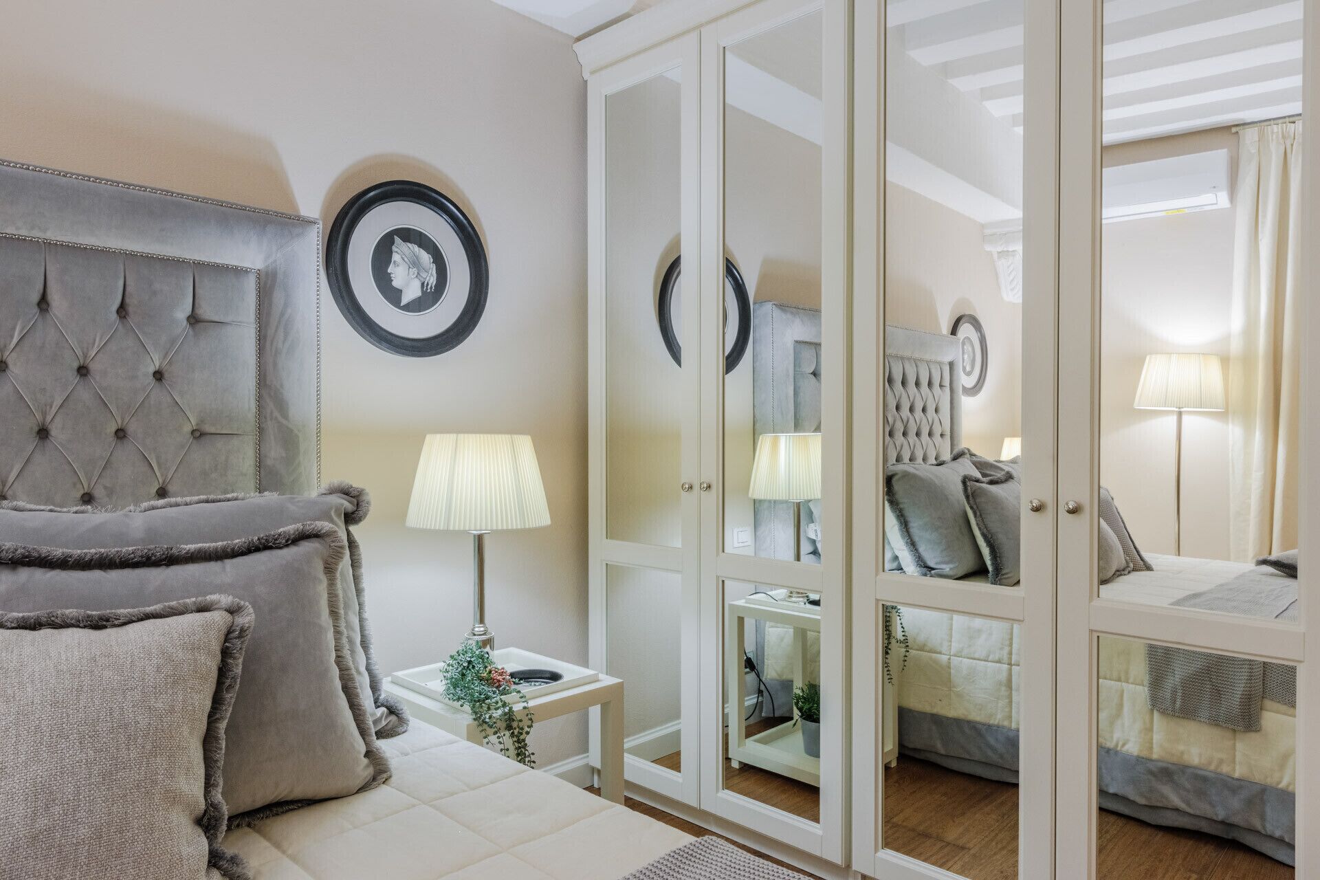 Camera da letto con testiera trapuntata, comodino con lampada, uno specchio e un letto con cuscini.